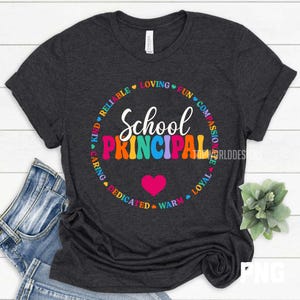 K&ouml;nnte beinhalten: Dunkelgraues T-Shirt mit den Worten "School Principal" in bunten Buchstaben. Ein Kreis aus W&ouml;rtern wie "Loving", "Fun" und "Loyal" umgibt den Text. Ein rosa Herz befindet sich darunter.