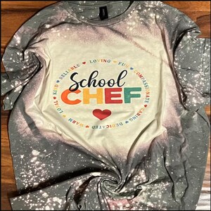 May include: A grey t-shirt with a bleach-splattered design. The shirt features a circle with the words "School Chef" in colourful letters and a heart. The circle is surrounded by the words "Reliable, Kind, Loyal, Warm, Dedicated, Caring, Compassionate, Fun, Loving".