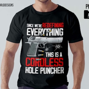 May include: Black t-shirt with a graphic of a silver handgun and bullets. Text reads "SINCE WE'RE REDEFINING EVERYTHING, THIS IS A CORDLESS HOLE PUNCHER." The shirt has a crew neck and short sleeves.