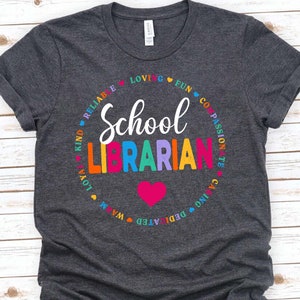 Op de afbeelding: Een donkergrijs T-shirt met een kleurrijk cirkelontwerp met de tekst "School Librarian" in een regenboogkleurig lettertype. De cirkel is omgeven door woorden als "Kind", "Reliable", "Loving", "Fun", "Compassionate", "Caring", "Dedicated", "Warm", "Loyal".