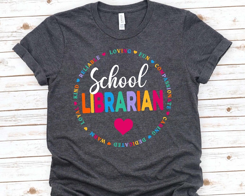 Op de afbeelding: Een donkergrijs T-shirt met een kleurrijk cirkelontwerp met de tekst "School Librarian" in een regenboogkleurig lettertype. De cirkel is omgeven door woorden als "Kind", "Reliable", "Loving", "Fun", "Compassionate", "Caring", "Dedicated", "Warm", "Loyal".