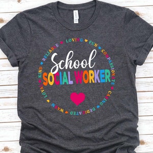 May include: A dark grey t-shirt with a circle design featuring the words "School Social Worker" in rainbow colors. The circle is surrounded by words describing positive qualities, such as "kind", "reliable", "loving", "fun", "compassionate", "caring", "dedicated", "warm", "loyal", and "passionate".