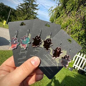 K&ouml;nnte beinhalten: Drei Paare geisterf&ouml;rmiger Ohrringe mit einer verspiegelten Oberfl&auml;che. Die Ohrringe befinden sich auf schwarzen Karten mit dem Text "HANDCRAFTED BY Jennifer Lynn" und einer Website-Adresse.