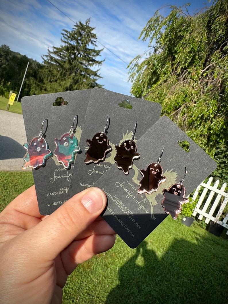 K&ouml;nnte beinhalten: Drei Paare geisterf&ouml;rmiger Ohrringe mit einer verspiegelten Oberfl&auml;che. Die Ohrringe befinden sich auf schwarzen Karten mit dem Text "HANDCRAFTED BY Jennifer Lynn" und einer Website-Adresse.