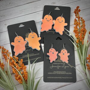 K&ouml;nnte beinhalten: Drei Paare orangefarbener Geisterohrringe auf schwarzem Karton. Die Ohrringe sind handgefertigt und haben silberne Haken. Der Karton tr&auml;gt den Text "HANDCRAFTED BY Jennifer Lynn" und Website-Informationen.