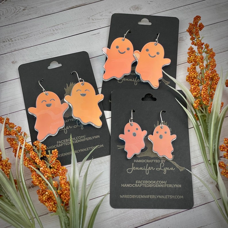 K&ouml;nnte beinhalten: Drei Paare orangefarbener Geisterohrringe auf schwarzem Karton. Die Ohrringe sind handgefertigt und haben silberne Haken. Der Karton tr&auml;gt den Text "HANDCRAFTED BY Jennifer Lynn" und Website-Informationen.