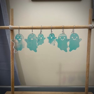 K&ouml;nnte beinhalten: Ein Set mit sechs transparenten Acryl-Geisterohrringen mit einem l&auml;chelnden Gesichtsdesign. Die Ohrringe h&auml;ngen an einem Holzst&auml;nder.
