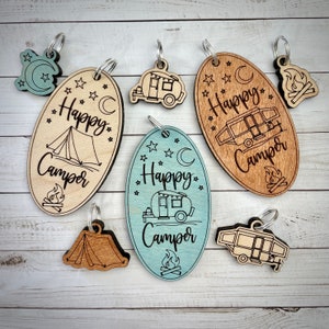 May include: Set of eight wooden keychains with camping-themed designs. The keychains feature a variety of designs, including a tent, a camper van, a campfire, and a crescent moon. The keychains are all made of natural wood and have a rustic look.