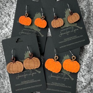 May include: Four pairs of pumpkin-shaped earrings on black cards. The earrings are made of wood and have a natural wood finish. The cards have the text "HANDCRAFTED BY Jennifer Lynn" and website information.