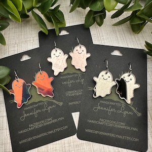 K&ouml;nnte beinhalten: Ein Set mit vier Paaren Halloween-geister-Ohrringen. Die Ohrringe sind aus klarem Acryl und haben ein Geisterdesign mit einem freundlichen Gesicht. Die Geister sind in verschiedenen Farben bemalt, darunter Orange, Pink und Silber. Die Ohrringe sind handgefertigt und werden mit einer schwarzen Karte geliefert, die den Namen des K&uuml;nstlers und seine Website enth&auml;lt.