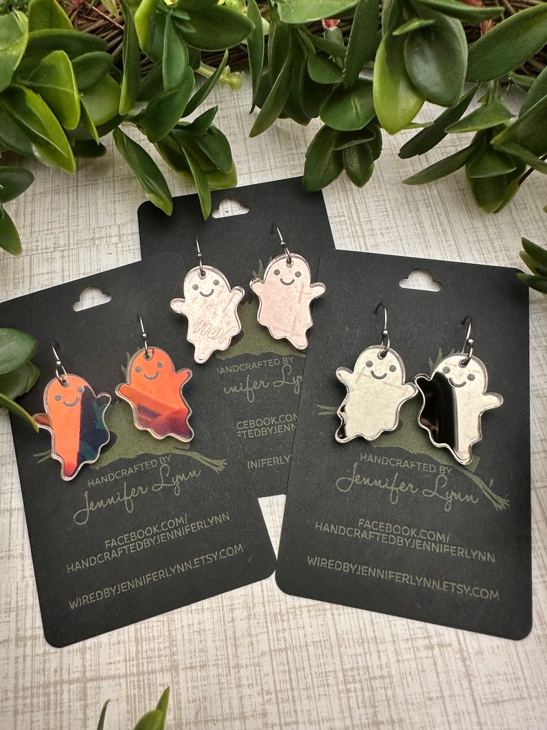 K&ouml;nnte beinhalten: Ein Set mit vier Paaren Halloween-geister-Ohrringen. Die Ohrringe sind aus klarem Acryl und haben ein Geisterdesign mit einem freundlichen Gesicht. Die Geister sind in verschiedenen Farben bemalt, darunter Orange, Pink und Silber. Die Ohrringe sind handgefertigt und werden mit einer schwarzen Karte geliefert, die den Namen des K&uuml;nstlers und seine Website enth&auml;lt.