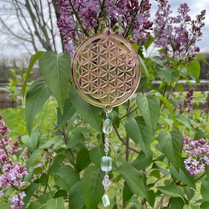 May include: A rose gold metal flower of life ornament with a crystal prism and beads hanging from it. The ornament is hanging from a lilac bush.