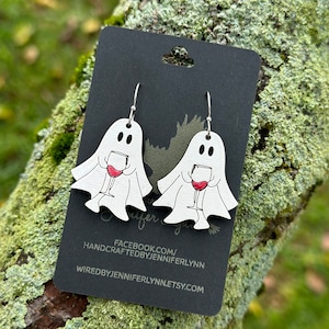 May include: A pair of white ghost earrings with red wine glasses. The earrings are on a black card with the text "FACEBOOK.COM/HANDCRAFTEDBYJENNIFERLYNN WIREDBYJENNIFERLYNN.ETSY.COM".