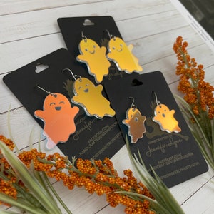 K&ouml;nnte beinhalten: Ein Set mit vier regenbogenfarbenen Geister-Ohrringen. Die Ohrringe sind aus Acryl und haben eine spiegelnde Oberfl&auml;che. Die Geister l&auml;cheln und haben einen gruseligen Ausdruck. Die Ohrringe sind handgefertigt und werden auf schwarzem Karton mit dem Namen des K&uuml;nstlers und seinen Social-Media-Informationen geliefert.