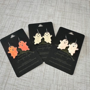 K&ouml;nnte beinhalten: Drei Paare Geister-Ohrringe auf schwarzem Karton. Die Ohrringe sind aus klarem Acryl mit einem wei&szlig;en, orangen und pfirsichfarbenen Glitzerfinish. Die Karten tragen den Text "HANDCRAFTED BY Jennifer Lynn" und Social-Media-Informationen.