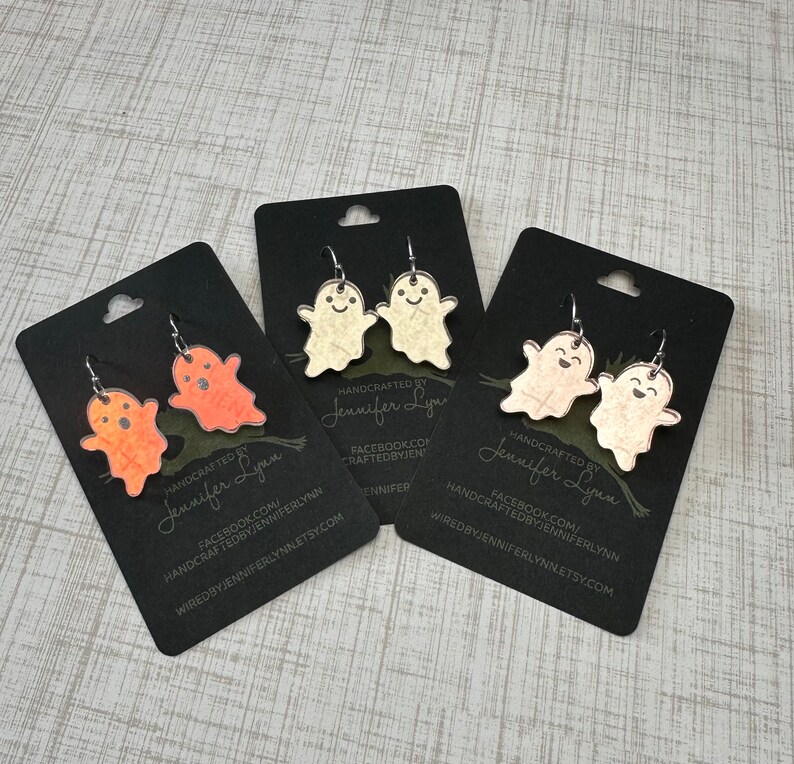 K&ouml;nnte beinhalten: Drei Paare Geister-Ohrringe auf schwarzem Karton. Die Ohrringe sind aus klarem Acryl mit einem wei&szlig;en, orangen und pfirsichfarbenen Glitzerfinish. Die Karten tragen den Text "HANDCRAFTED BY Jennifer Lynn" und Social-Media-Informationen.