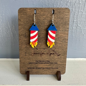 May include: A pair of red, white, and blue firework-shaped earrings on a wooden display stand. The earrings are made of wood and have silver hooks. The stand has the text "Jennifer Lynn" and contact information for the seller.