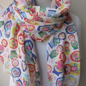 May include: White scarf with a repeating pattern of colorful Russian nesting dolls. The dolls are arranged in a random pattern on a white background with small pink hearts.