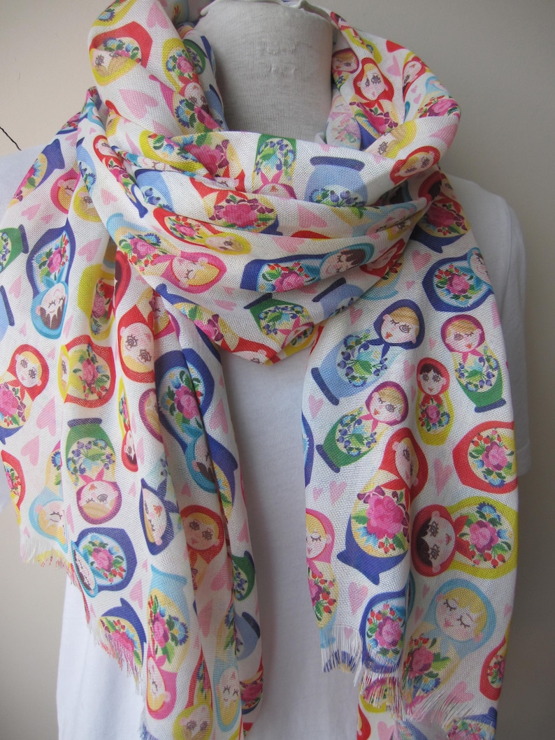 May include: White scarf with a repeating pattern of colorful Russian nesting dolls. The dolls are arranged in a random pattern on a white background with small pink hearts.