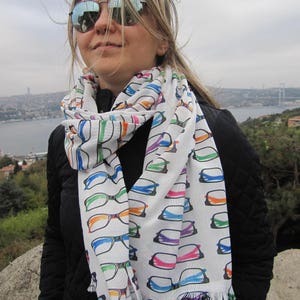 May include: A woman wearing a black jacket and a white scarf with a colorful eyeglasses print. The scarf has a fringed edge.