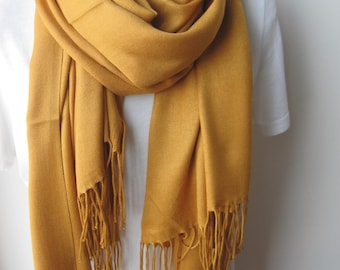 May include: A mustard yellow pashmina scarf with fringe. The scarf is draped over a white shirt.
