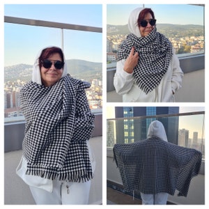 May include: A black and white houndstooth patterned shawl with fringe. The shawl is draped over a person wearing a white hooded sweatshirt and sunglasses. The background shows a cityscape and a clear sky.