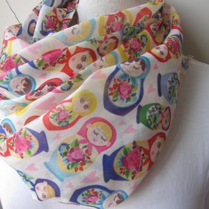 May include: A white scarf with a colorful pattern of Russian nesting dolls. The dolls are depicted in various sizes and colors, with floral designs and intricate details.