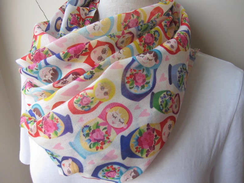 May include: A white scarf with a colorful pattern of Russian nesting dolls. The dolls are depicted in various sizes and colors, with floral designs and intricate details.