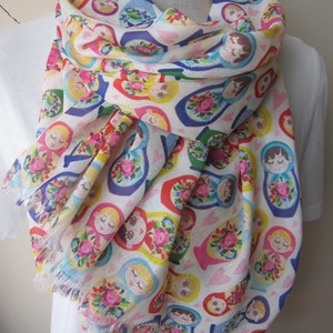 May include: A white scarf with a colorful pattern of Russian nesting dolls. The dolls are all different sizes and colors, and they are arranged in a random pattern. The scarf has a fringed edge.