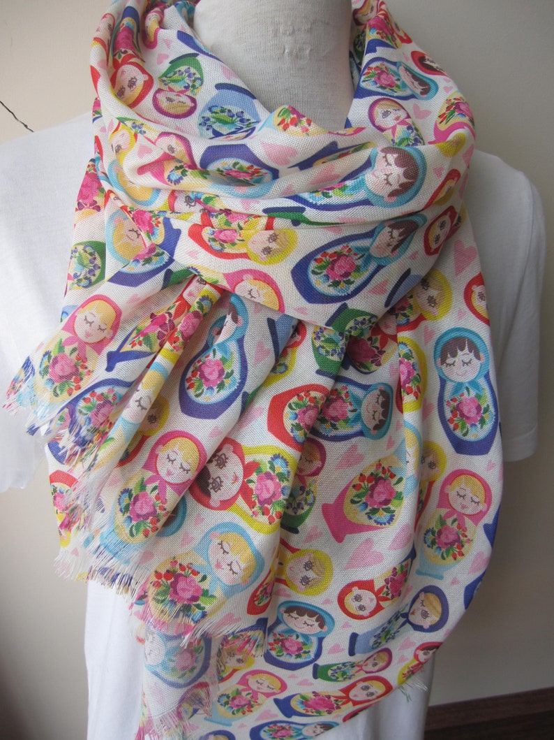 May include: A white scarf with a colorful pattern of Russian nesting dolls. The dolls are all different sizes and colors, and they are arranged in a random pattern. The scarf has a fringed edge.