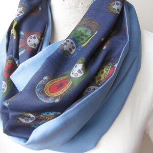 May include: A blue infinity scarf with a patterned interior featuring colorful doll-like figures. The scarf has a light blue border and is draped around a mannequin's neck, showcasing its design and texture.