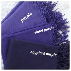May include: Three purple fabric scarves with fringed edges. The scarves are layered on top of each other, with the lightest shade of purple on top and the darkest shade on the bottom. The scarves are labeled "purple", "violet purple", and "eggplant purple".