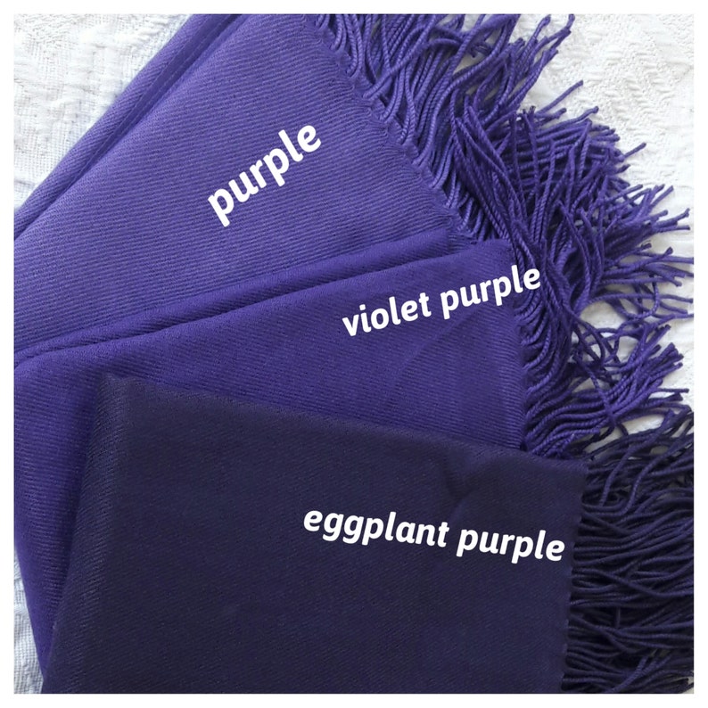 May include: Three purple fabric scarves with fringed edges. The scarves are layered on top of each other, with the lightest shade of purple on top and the darkest shade on the bottom. The scarves are labeled "purple", "violet purple", and "eggplant purple".