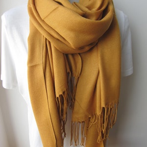 May include: A mustard yellow pashmina scarf with fringed edges. The scarf is draped around a white shirt.