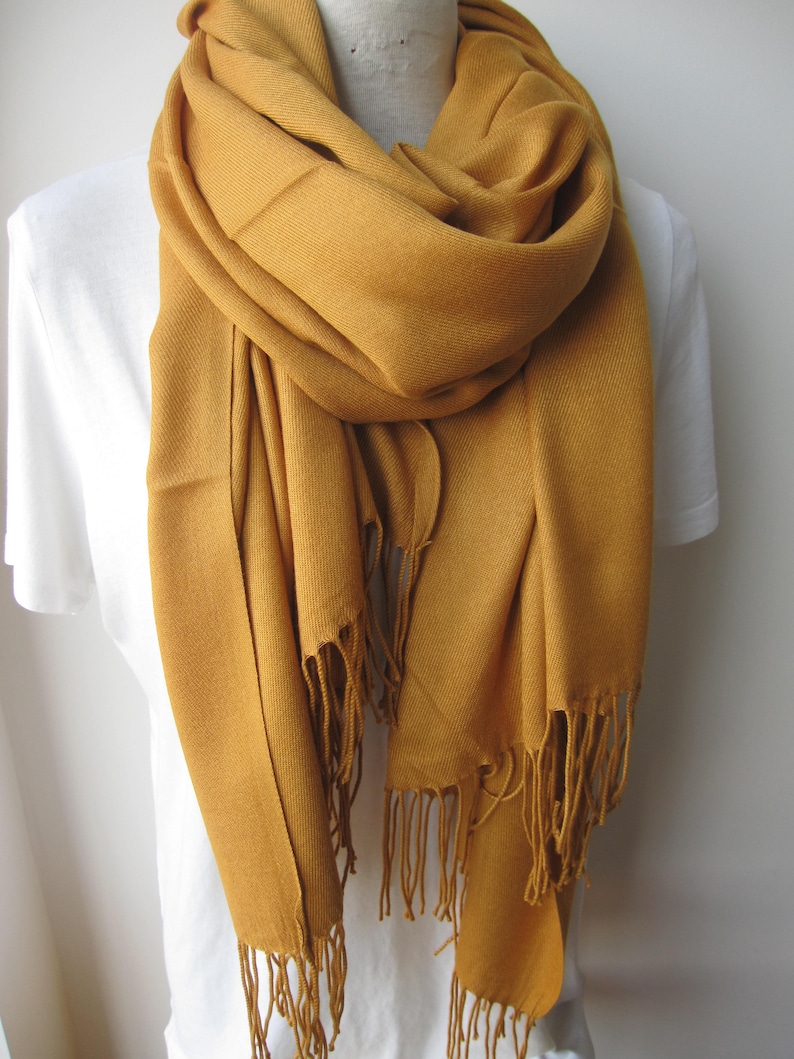 May include: A mustard yellow pashmina scarf with fringed edges. The scarf is draped around a white shirt.