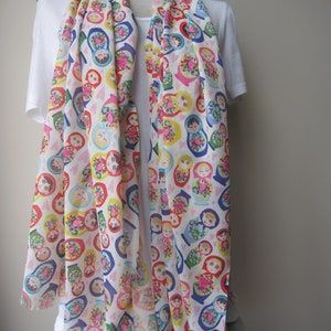 May include: A white scarf with a colorful pattern of Russian nesting dolls. The dolls are depicted in various sizes and colors, with each doll wearing a different traditional dress. The scarf has a fringed edge.