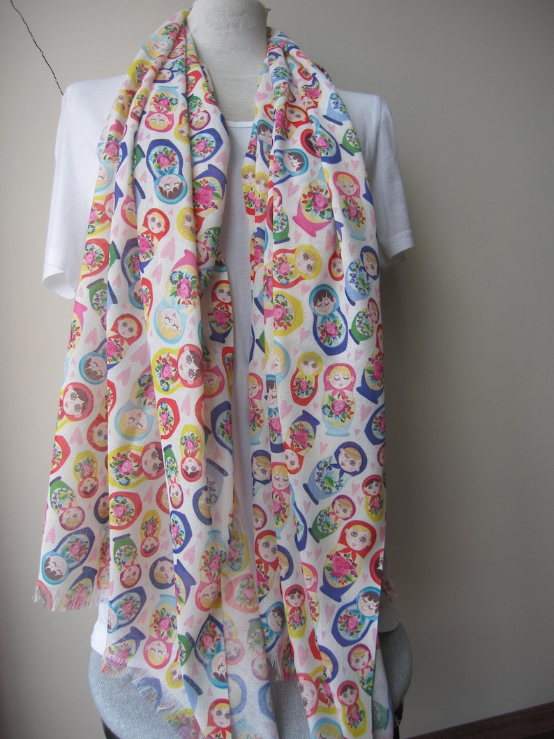 May include: A white scarf with a colorful pattern of Russian nesting dolls. The dolls are depicted in various sizes and colors, with each doll wearing a different traditional dress. The scarf has a fringed edge.