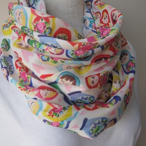 May include: A white infinity scarf with a colorful pattern of Russian nesting dolls. The dolls are depicted in various sizes and colors, with floral accents.