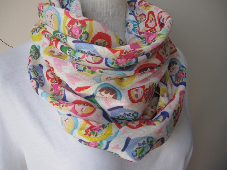May include: A white infinity scarf with a colorful pattern of Russian nesting dolls. The dolls are depicted in various sizes and colors, with floral accents.