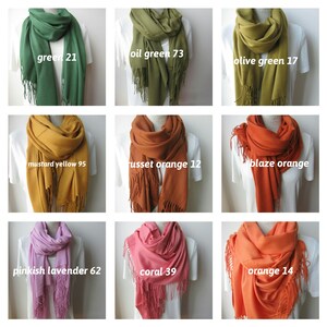 May include: Nine different colors of scarves with fringe. The colors are green, oil green, olive green, mustard yellow, russet orange, blaze orange, pinkish lavender, coral, and orange. Each scarf is draped over a white shirt on a mannequin.