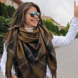 May include: A woman wearing a brown and black plaid scarf with fringe. She is wearing a white long-sleeved shirt and sunglasses.