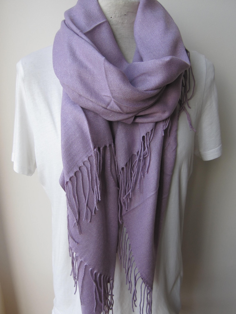 May include: A soft, lavender-colored scarf with fringed edges. The scarf is draped around a white shirt.