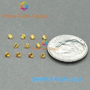 May include: A collection of small, round, yellow gemstones arranged on a gray surface. A U.S. dime is included for size comparison. The gemstones are a vibrant yellow color. The text "Prime Gem Source" is visible in the image.