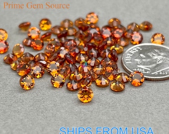 May include: A close-up image of a pile of small, round, orange-brown gemstones. The gemstones are scattered on a gray surface, and a US dime is included for scale.