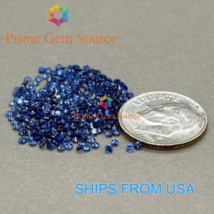 Op de afbeelding: Een verzameling kleine, gefacetteerde blauwe edelstenen op een grijze ondergrond, naast een Amerikaanse dime-munt. De edelstenen zijn diepblauw. De tekst "Prime Gem Source" en "SHIPS FROM USA" zijn zichtbaar.