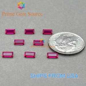 May include: A collection of rectangular, vibrant pink gemstones, possibly rubies, are displayed on a gray surface next to a US dime for size comparison. The gemstones are evenly spaced and reflect light, showcasing their polished surfaces.