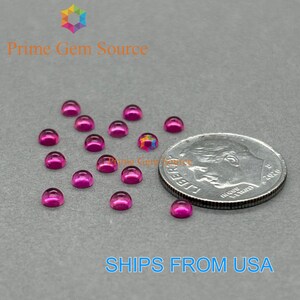 May include: A collection of small, vibrant magenta-colored gemstones with a domed, cabochon cut. A US dime is included for size reference. The gemstones are scattered on a gray surface. The text "Prime Gem Source" is visible.