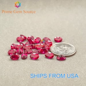 May include: A close-up of a pile of oval-shaped, red gemstones. The gemstones are scattered on a light gray surface. A US dime is included for scale.