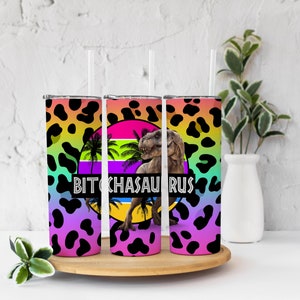 May include: Three stainless steel tumblers with lids and straws. The tumblers have a rainbow gradient background with a black leopard print overlay. The center tumbler has a graphic of a dinosaur with the text "BITCHASAURUS" in rainbow letters.
