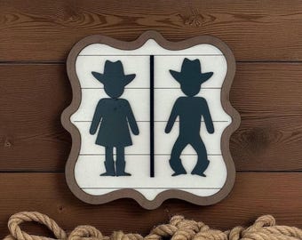 May include: A wooden restroom sign with a brown frame and a white background. The sign features silhouettes of a person wearing a cowboy hat and boots. The sign is mounted on a wooden wall.